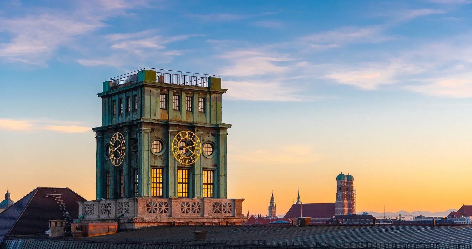 The Thiersch tower on the roof of the TUM main building at sunset, its clock faces lit, with the Frauenkirche beyond it across Munich